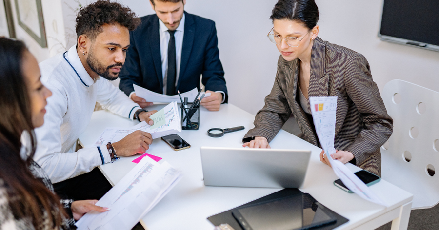 Four coworkers reviewing printed charts and graphs around a laptop during a business meeting.