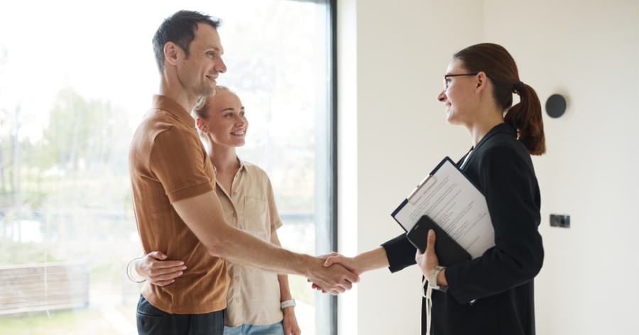 Real estate agent shaking hands with a smiling couple in a bright, window-lined room, holding a clipboard.