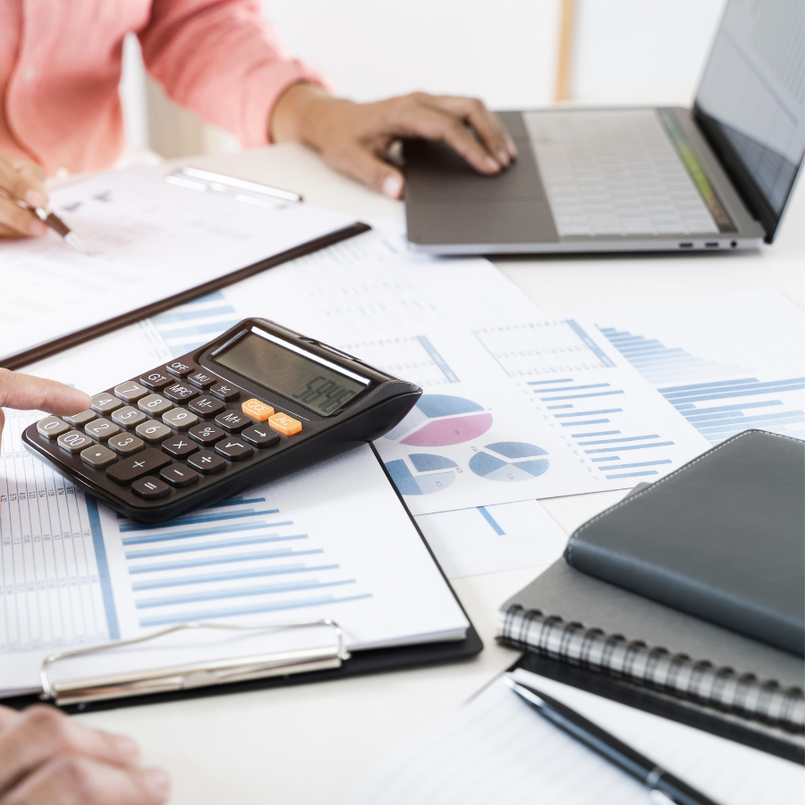 Hands using a calculator and laptop surrounded by financial charts and reports