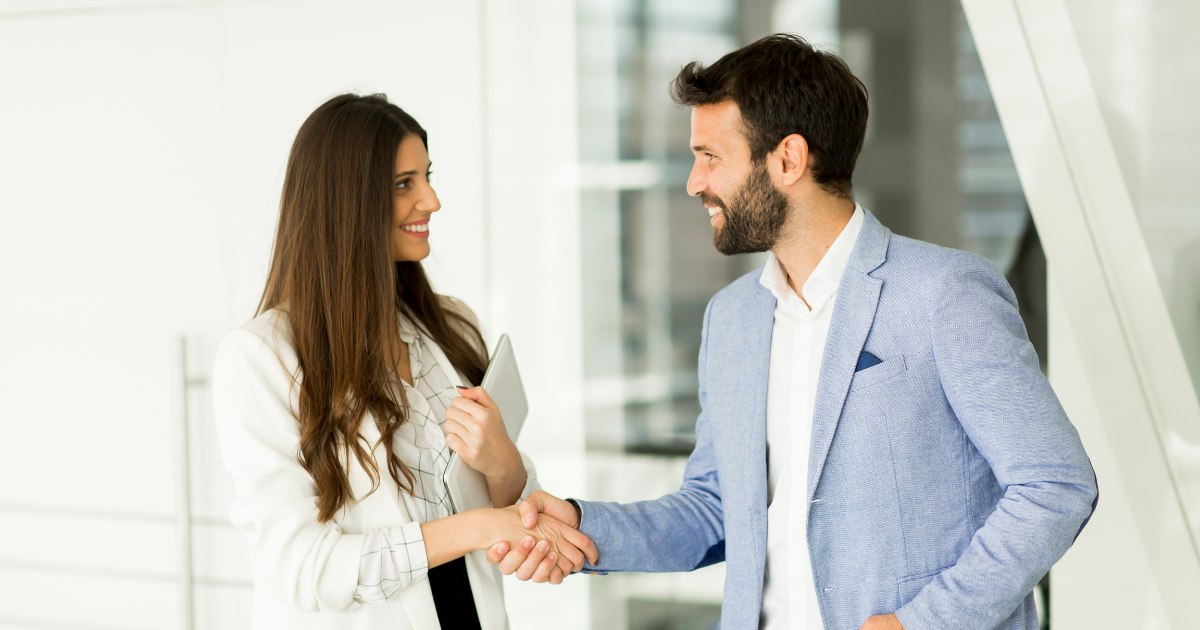 A woman and a man in business attire shaking hands and smiling in a modern office