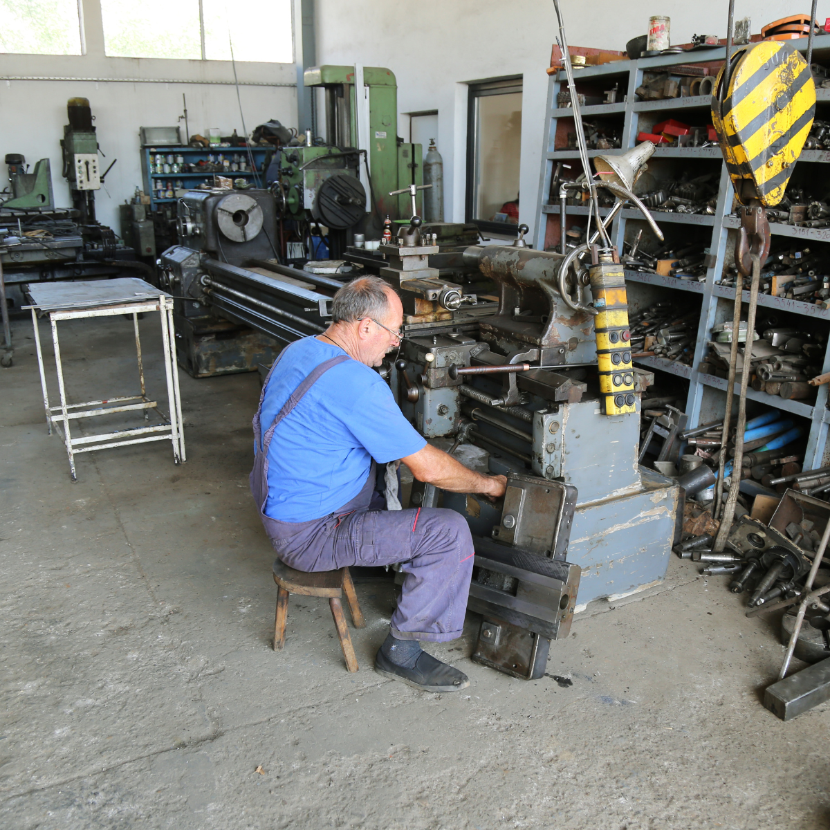  A middle-aged male machinist in blue coveralls seated on a stool while operating a large metal lathe in a cluttered workshop filled with tools, parts, and industrial equipment.