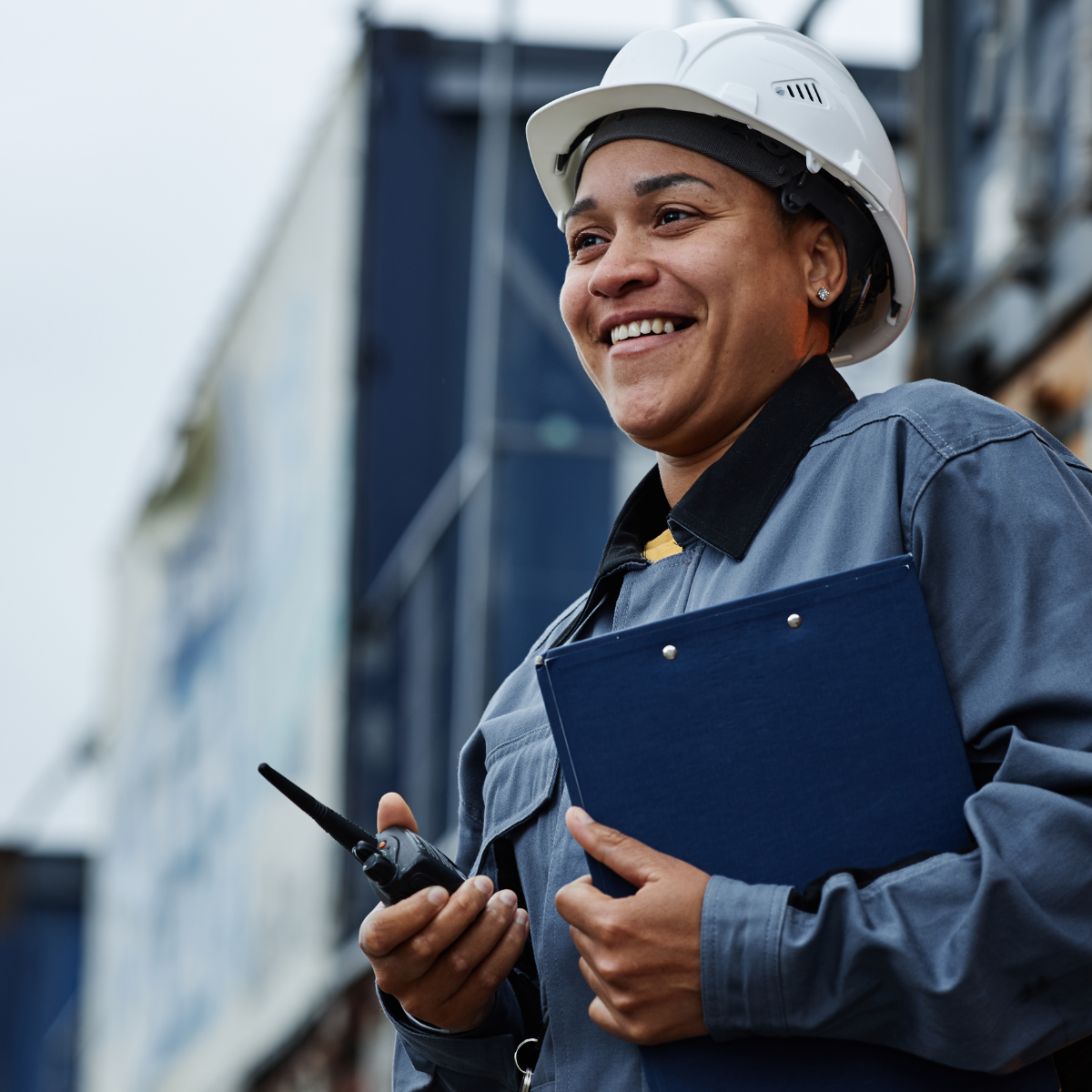 A smiling female industrial worker wearing a white hard hat and grey work jacket, holding a clipboard and walkie-talkie outdoors in front of a large industrial structure.