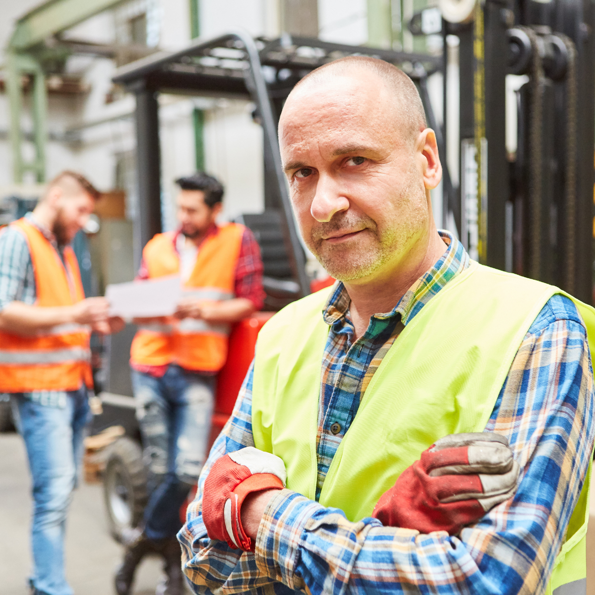 A confident male warehouse worker in a yellow safety vest and red work gloves standing with arms crossed in the foreground, with two colleagues in orange vests reviewing documents beside a forklift in the background.