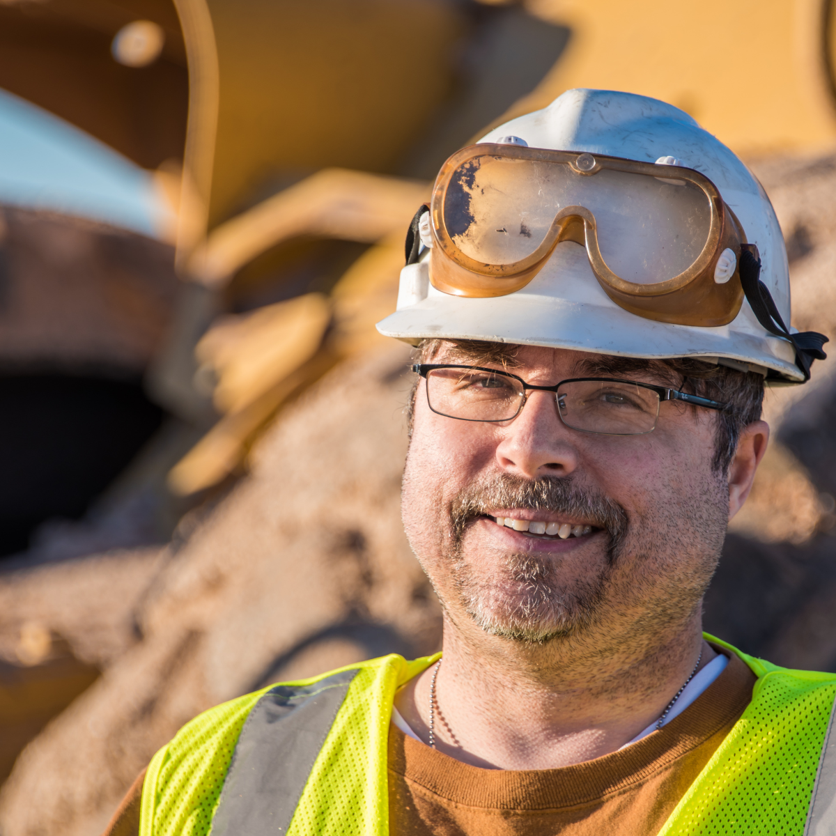 A smiling male construction worker wearing a white hard hat with safety goggles pushed up, glasses, and a yellow safety vest, with heavy yellow equipment visible in the background.